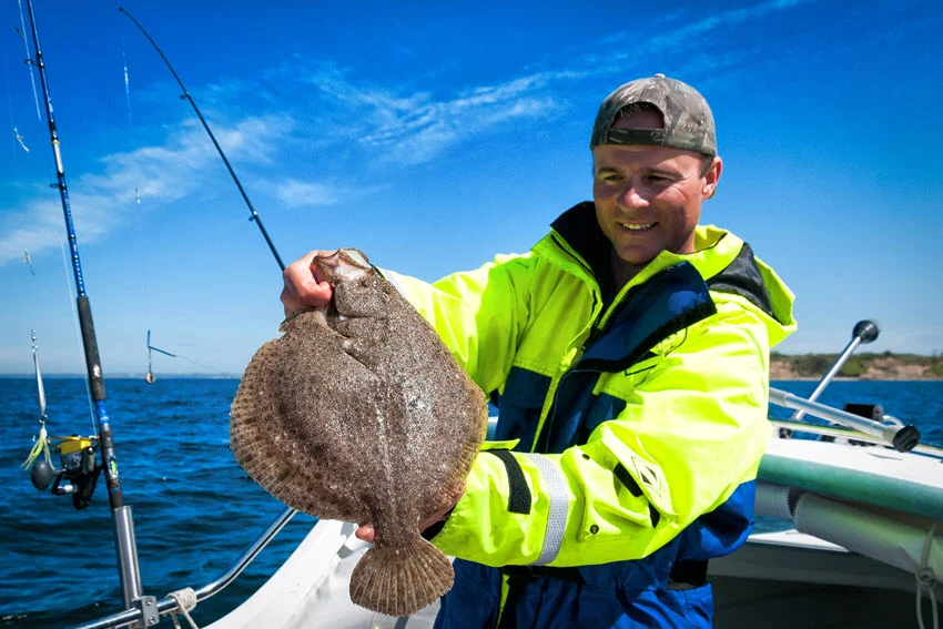 A smiling angler stands on a fishing boat, with jigging rods attached to the boat, on a bright sunny day, with a dark blue ocean in the background, holding freshly caught Flounder.