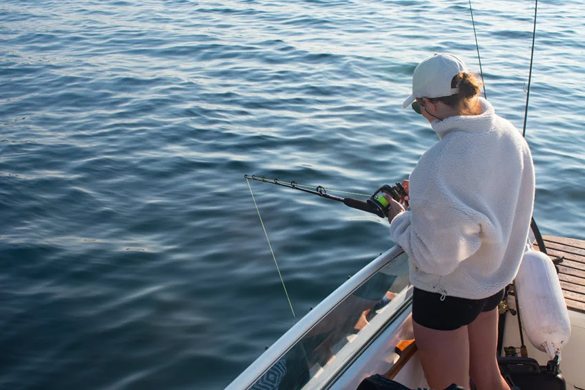 Back view of a female angler standing on the bow of a moving fishing boat, preparing a jigging rod and reel.