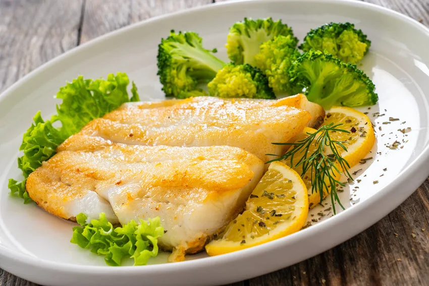 Seared halibut fillet, sliced lemon and boiled broccoli served in a white plate, and placed on wooden table.