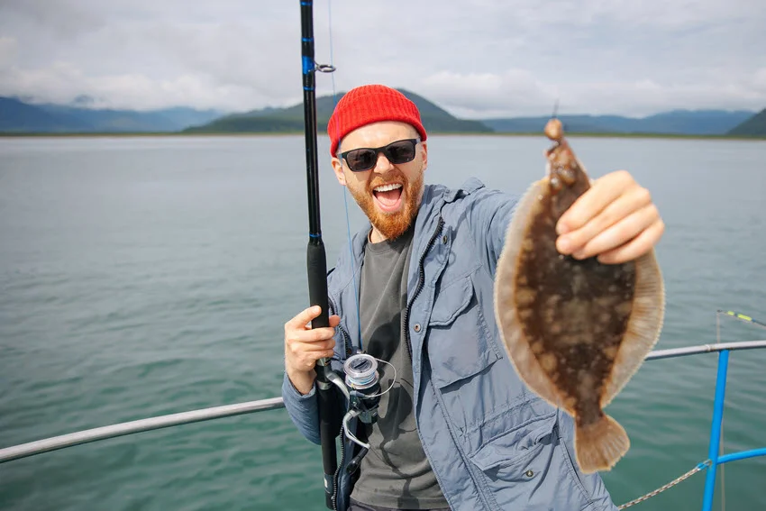 A happy, smiling angler stands on a boat and holds the rod in one and freshly caught Flounder in the other hand, with a blurry ocean in the background.