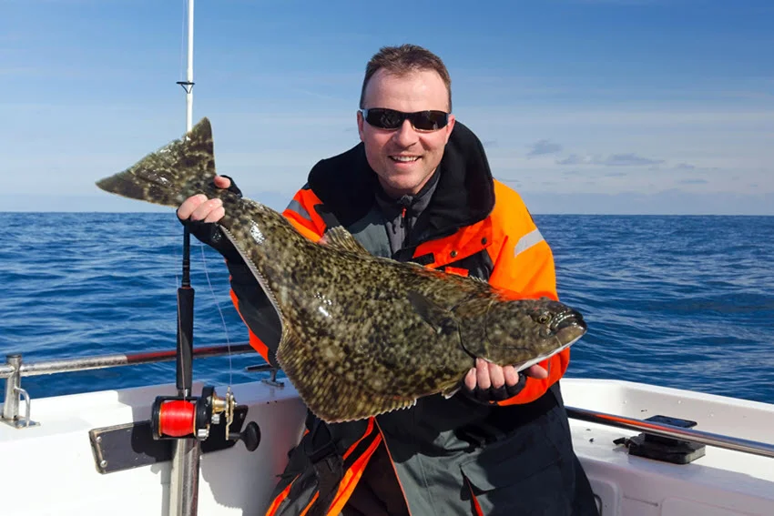 View of a happy angler wearing black sunglasses posing with a big Halibut caught on a boat.