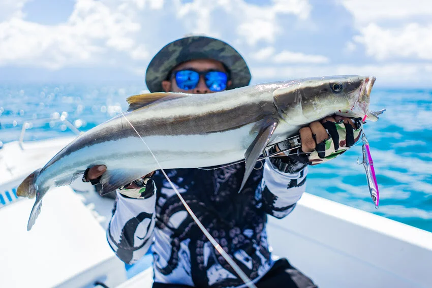 An angler, wearing polarized glasses and a hat, sits on a moving boat and holds a freshly caught Cobia with both hands.