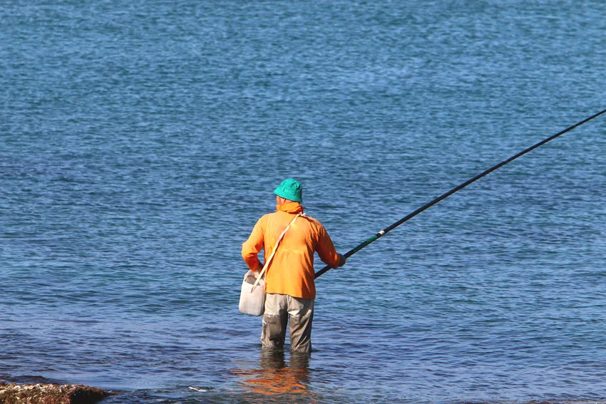 Back view of a wade angler holding a bucket and rod while standing in the shallows fishing.