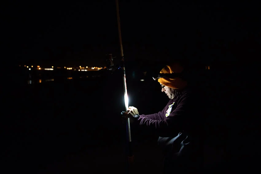 A side view of a lone angler in the dark, wearing a headlight and preparing their pole for night gig fishing, with the light casting a focused beam on their gear.