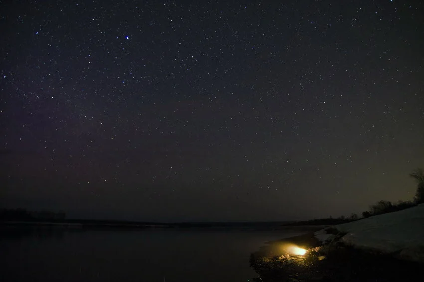  A panoramic view of a starry night sky above a calm river, with a warm, glowing campfire on the riverbank casting light on the surrounding area and creating a cozy ambiance.