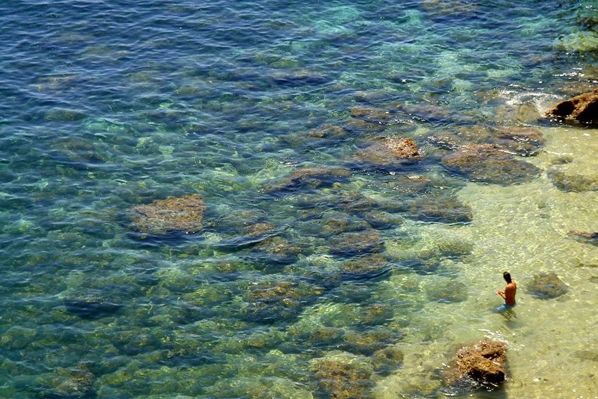 An aerial view of the shallow, crystal blue, and rocky sea coastline.