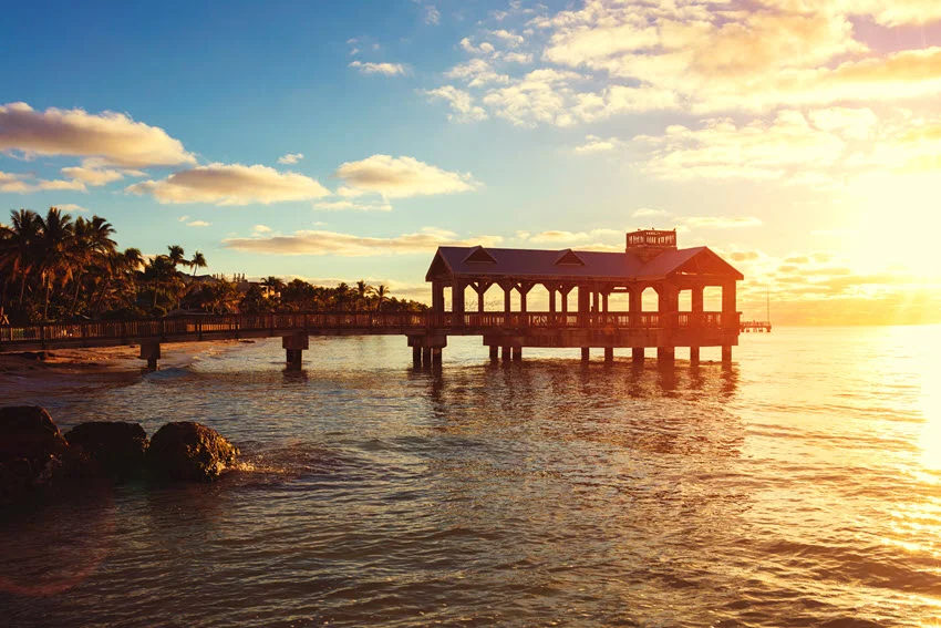 Sunrise view of a pier in Key West, Florida, with calm water, warm golden light, and tropical shoreline scenery.