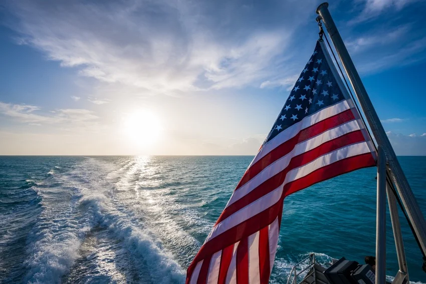 An American flag flies from a boat at sunrise over the Gulf of Mexico, with open water and a foamy wake behind.