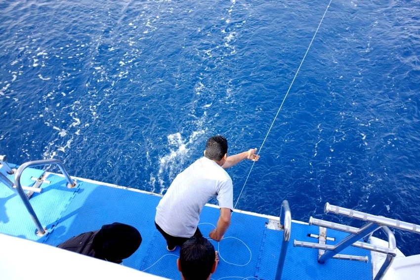 An angler fishes from the stern of a boat in deep blue water while another person watches from the deck.