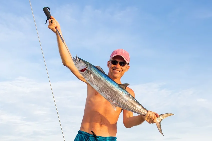 An angler holds a large Wahoo with blue vertical bars, a streamlined body, and a deeply forked tail.