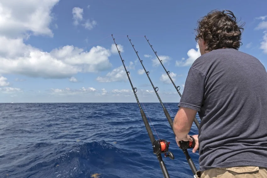 An angler stands beside three fishing rods on a boat, overlooking open water during an offshore fishing trip.