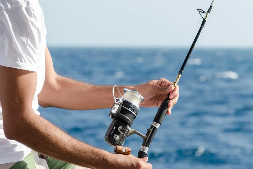 An angler holds a fishing rod fitted with a spinning reel, with the open sea visible in the background.