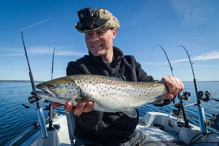 An angler with a smile stands on a trolling fishing boat, holding a giant Trout caught in the lake.