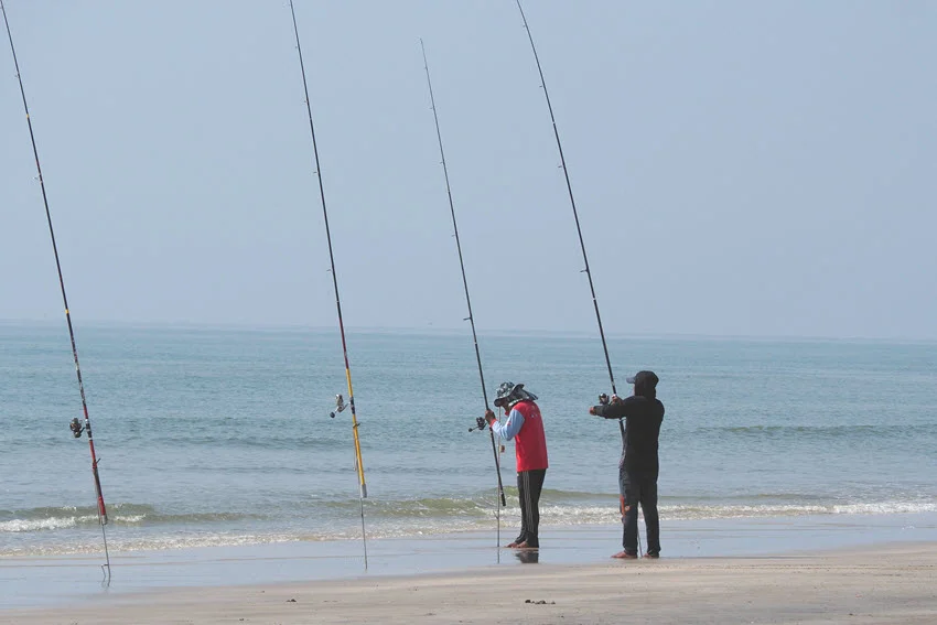 Distant view of two anglers standing on a beach, preparing their fishing rods for surf fishing.