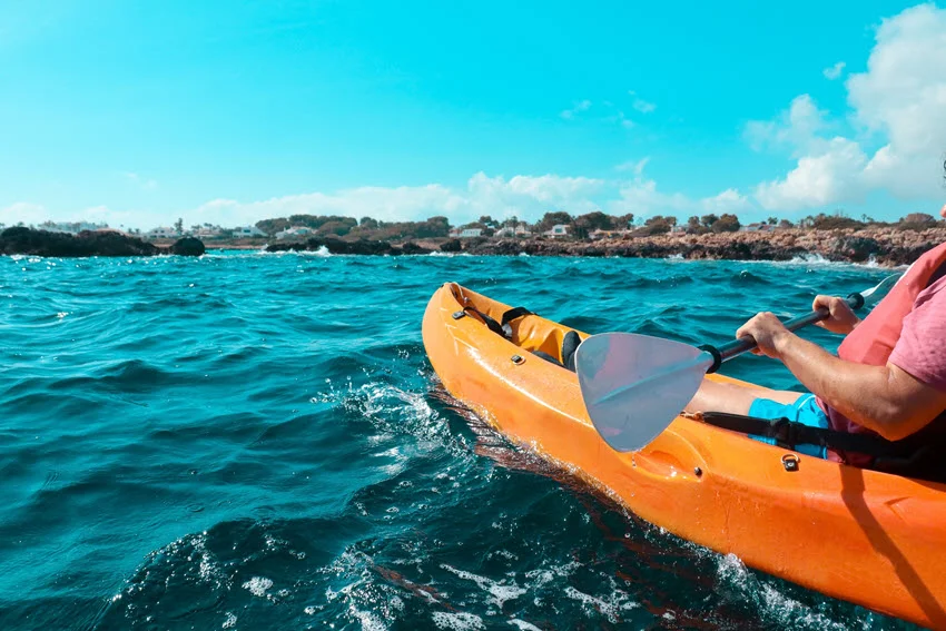 An angler in a bright orange kayak paddling through calm offshore waters under a clear sky.