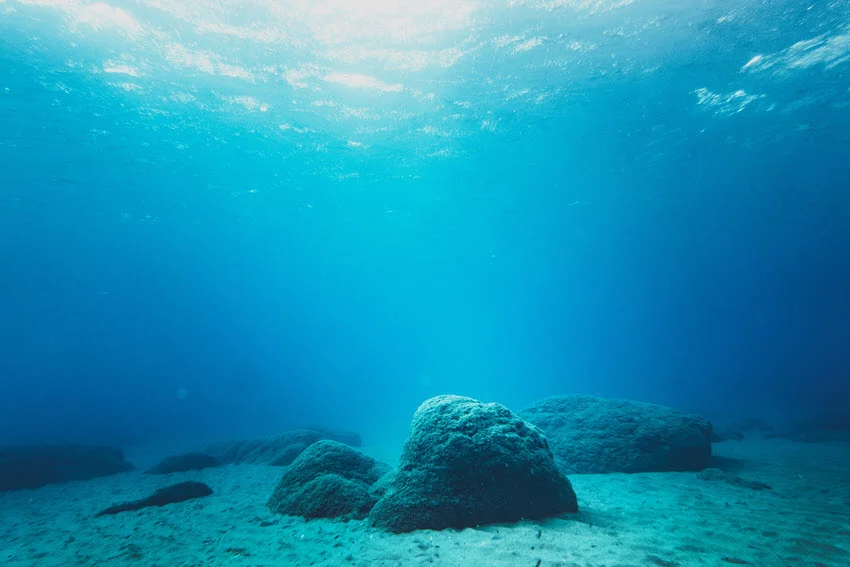 Underwater view of rock formations on the sandy ocean floor with sunlight rays streaming down from the surface.