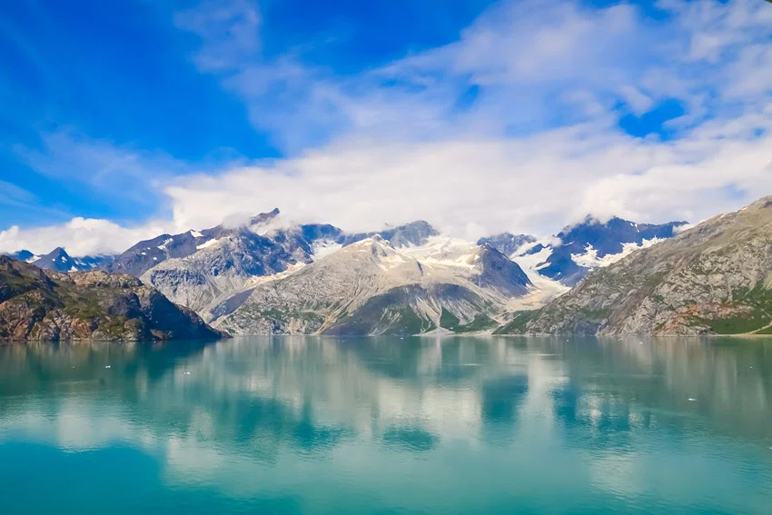 Snow-streaked mountains rise above calm turquoise water in Alaska, with rocky slopes and low clouds reflected beneath a bright blue sky.