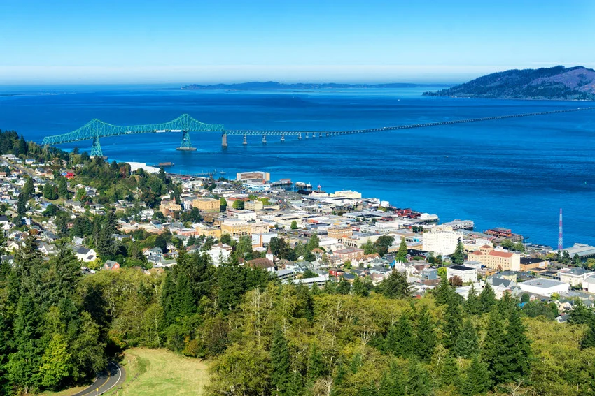 Astoria, Oregon, beside the Columbia River, with the Astoria-Megler Bridge stretching toward Washington beyond waterfront buildings and tree-covered slopes.