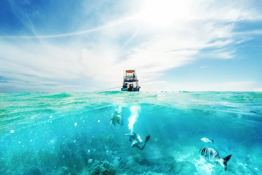 Scuba divers swimming among fish beneath a tour boat near Cozumel, Mexico, with a split-level view showing the sea surface and underwater terrain.