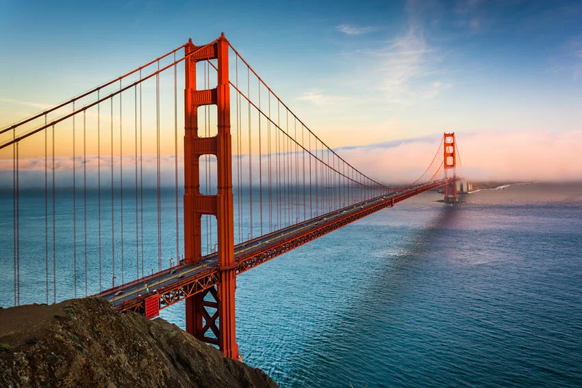 Golden Gate Bridge in San Francisco, California, at sunset, with fog around the far tower and blue water beneath its orange span.