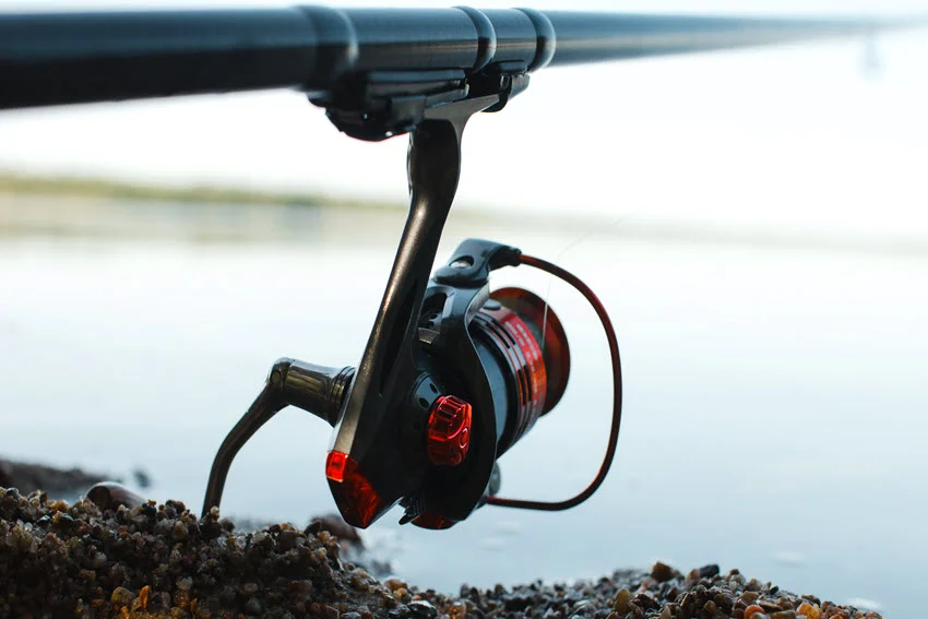 A black and red spinning reel mounted on a fishing rod rests on a pebbled shore beside calm water.