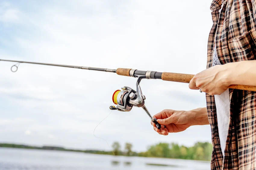 Angler holding a spinning rod beside calm water, used as a general illustration for jigging, with one hand on the reel handle.