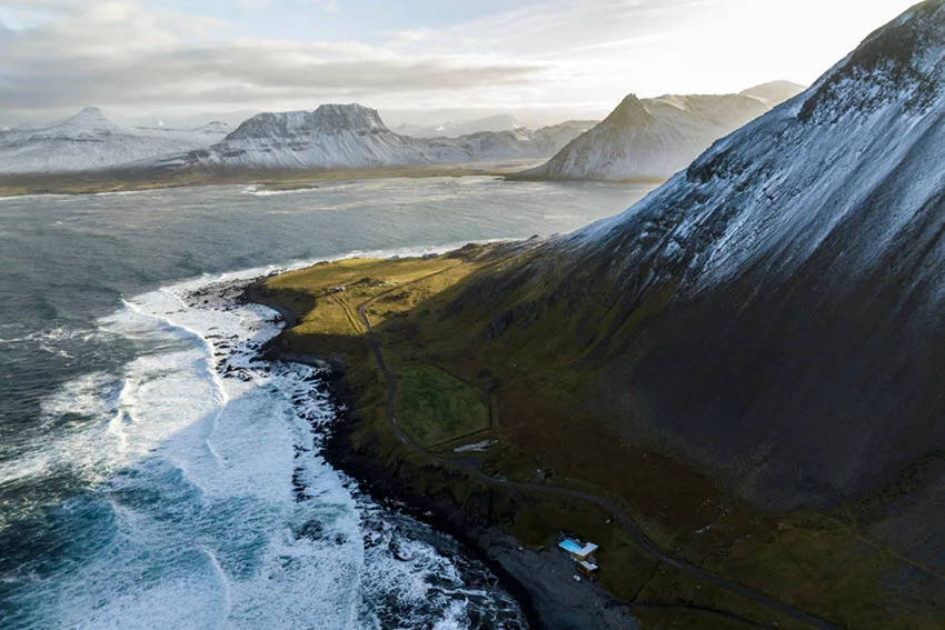 Snow-dusted mountains above Iceland's north coast and the Greenland Sea, with breaking waves, coastal grassland, and a narrow shoreline road.