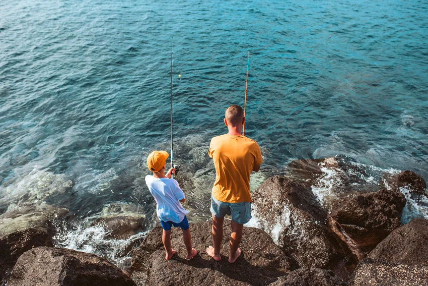 Two anglers fishing from coastal rocks, with submerged formations visible through clear water, illustrating rocky structure discussed in the fishing techniques section.