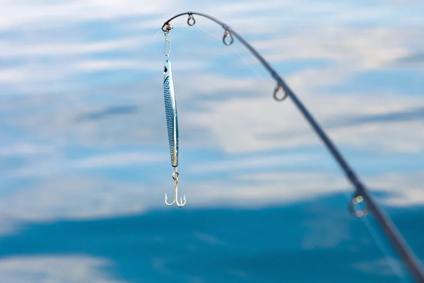 Slender silver metal lure with a treble hook hanging from a rod above blue water, illustrating artificial lures discussed for Halibut fishing.