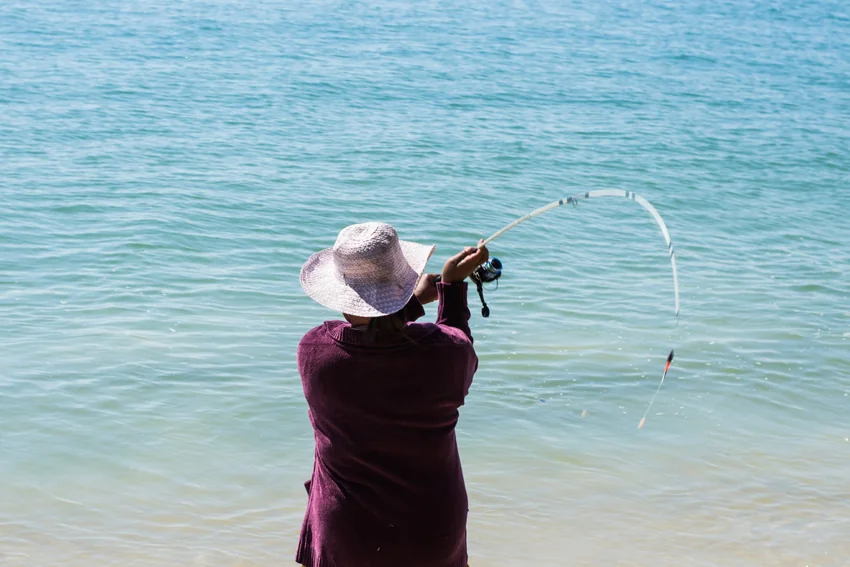 Angler wearing a sun hat casts a spinning rod beside the water, with the bent rod and suspended rig visible during the motion.