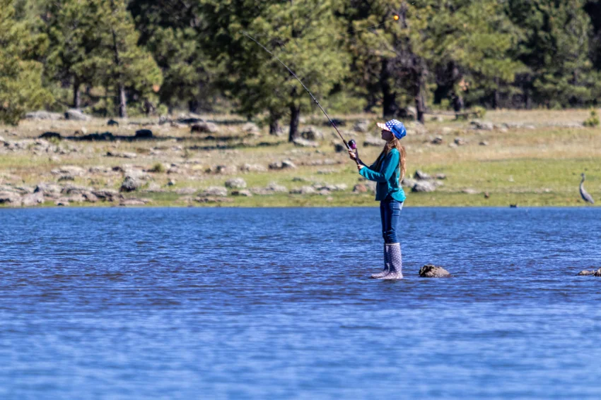 View of an angler wearing a blue cap holds a raised fishing rod in shallow water, with a float visible above and trees along the shoreline.