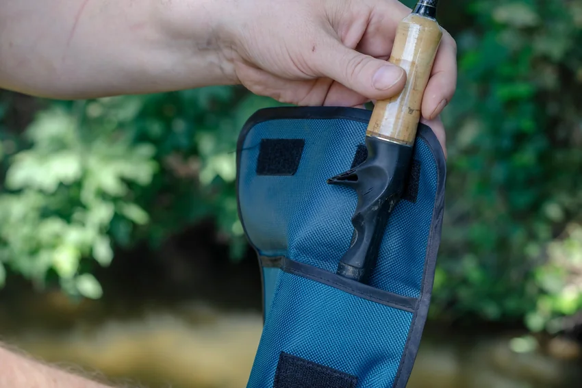 Hand holds a fishing rod with a cork handle and trigger grip beside an open blue protective case, against a leafy background.