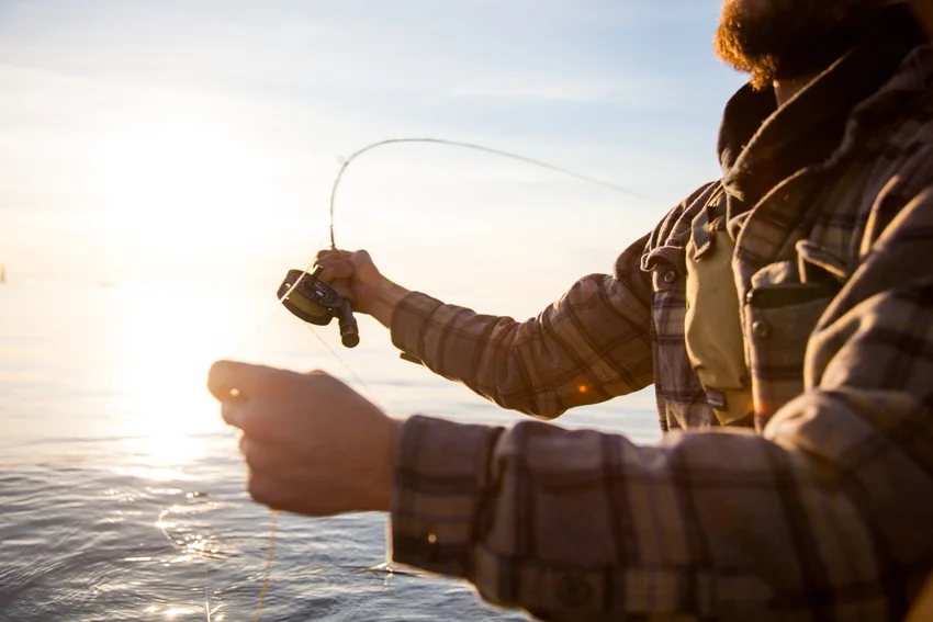 Angler wearing a plaid shirt holds a fly rod and reel while controlling loose line with the other hand above sunlit water.