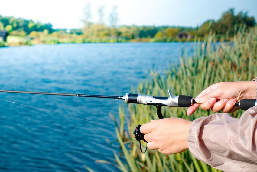Angler holds a spinning rod beside the water, with both hands, the reel seat, and part of the reel visible in the foreground.
