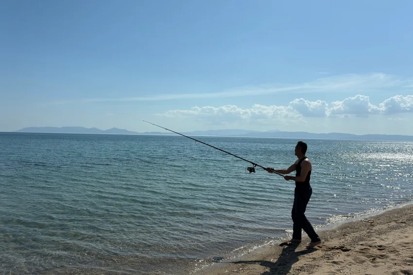Angler holds a spinning rod along an open shoreline, with gentle waves, a sandy bank, and distant hills visible beneath a blue sky.
