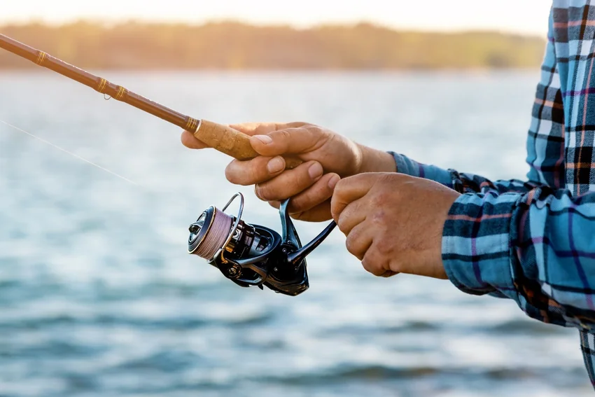 Angler holds a cork-handled rod and operates a spinning reel beside the water, with the spool, bail, and reel handle clearly visible.