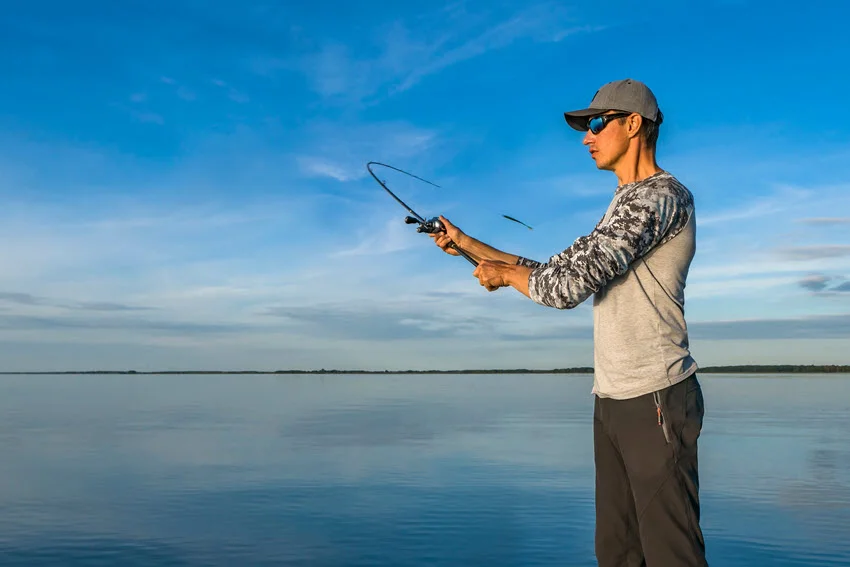 A side view of an angler wearing sunglasses and a gray jacket, casting a fishing rod with a clear sky and a forested shoreline in the distance.