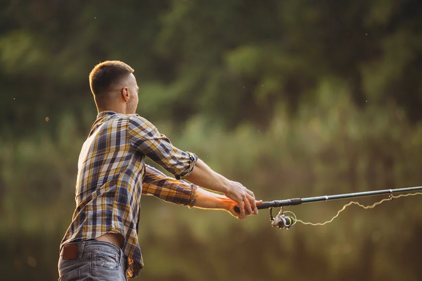 Back view of an angler dressed casually, casting a line into a river surrounded by dense vegetation.