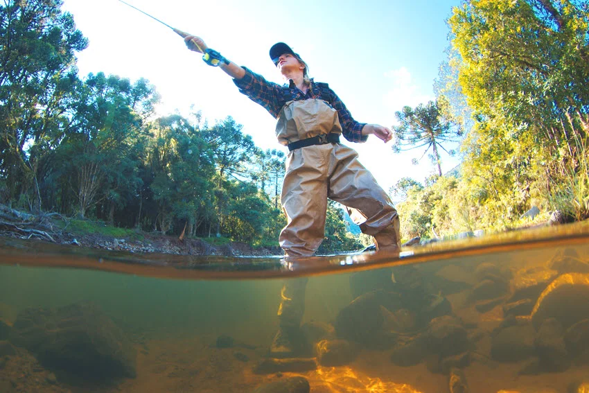 A view of a female angler casting her lure while standing in a river, dressed in wading gear and surrounded by trees.