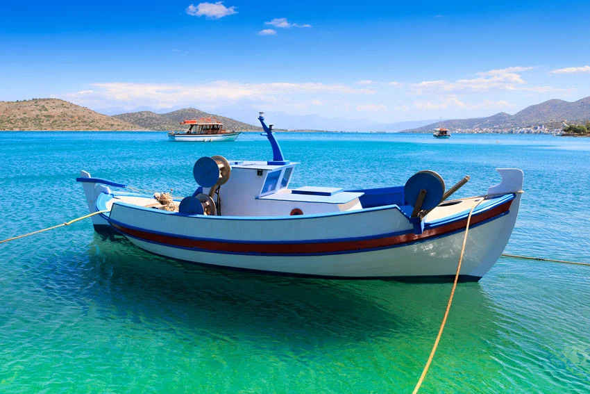 A small fishing boat anchored in a bay with blue-green water, mountains and other boats visible in the background.