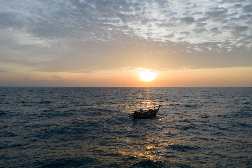 A view of a small fishing boat drifting in inshore waters, with a sunset in the distance reflecting off the choppy waves.