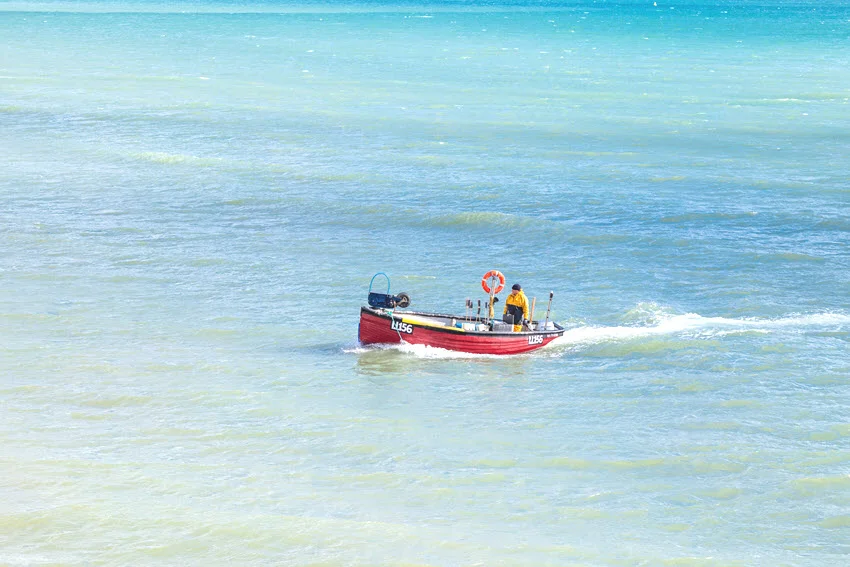 A distant view of a small red fishing boat with an angler in a yellow jacket, floating near the ocean shallows.