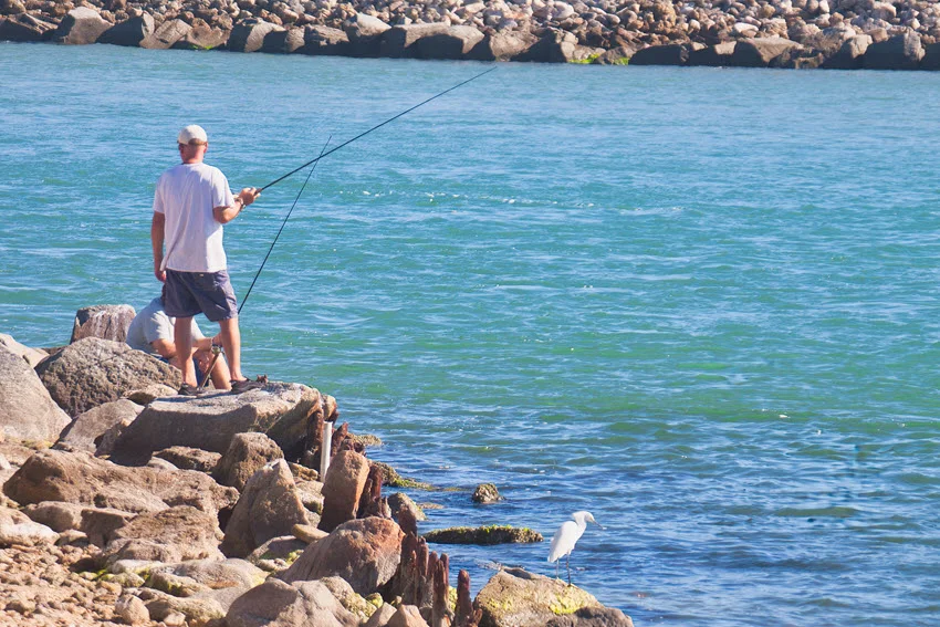 Angler casting from a rocky shoreline into clear saltwater near a coastal inlet.