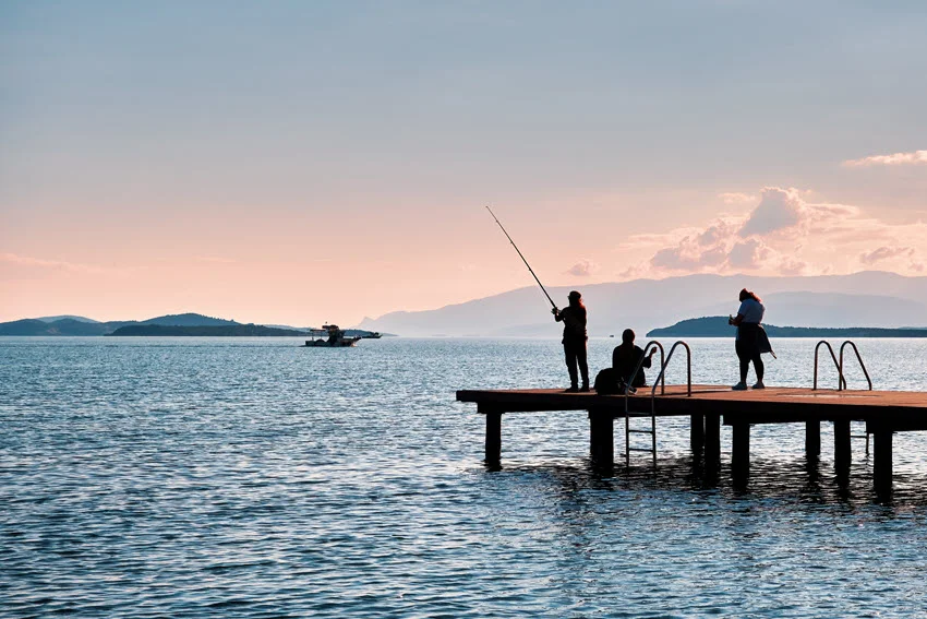 Silhouette view from a distance of several anglers standing on a fishing pier, performing kite fishing against the open sky.