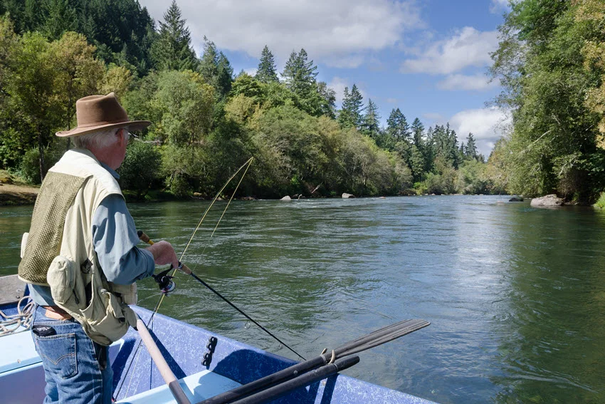 Back view of an elderly angler standing in a small drifting boat, holding a kite fishing rod and gazing out over the water.