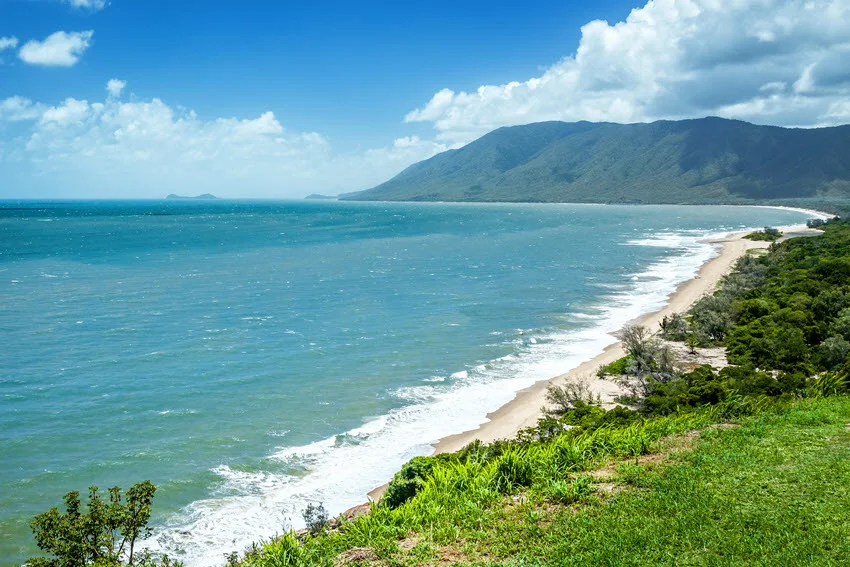 View of a beautiful beach in Queensland, Australia, with clear turquoise waters, soft sandy shores, and lush greenery in the background.
