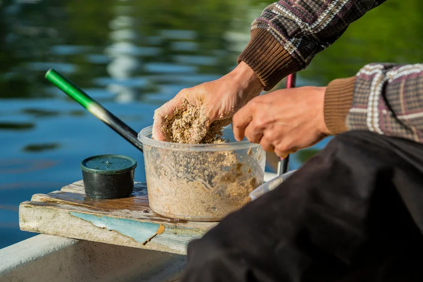Hands mixing bait in a plastic bowl to prepare fish food and fishing chum while sitting on a wooden board in a white boat.