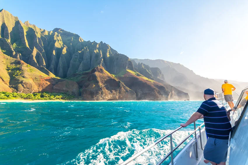 Boat tour with tourists cruising along the stunning Na Pali Coast at sunset on Kauai Island, Hawaii, with dramatic cliffs and golden skies.
