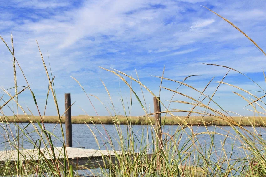 A sunny day with blue skies and soft clouds, showcasing a wooden dock extending over a peaceful river in the Biloxi Marsh.
