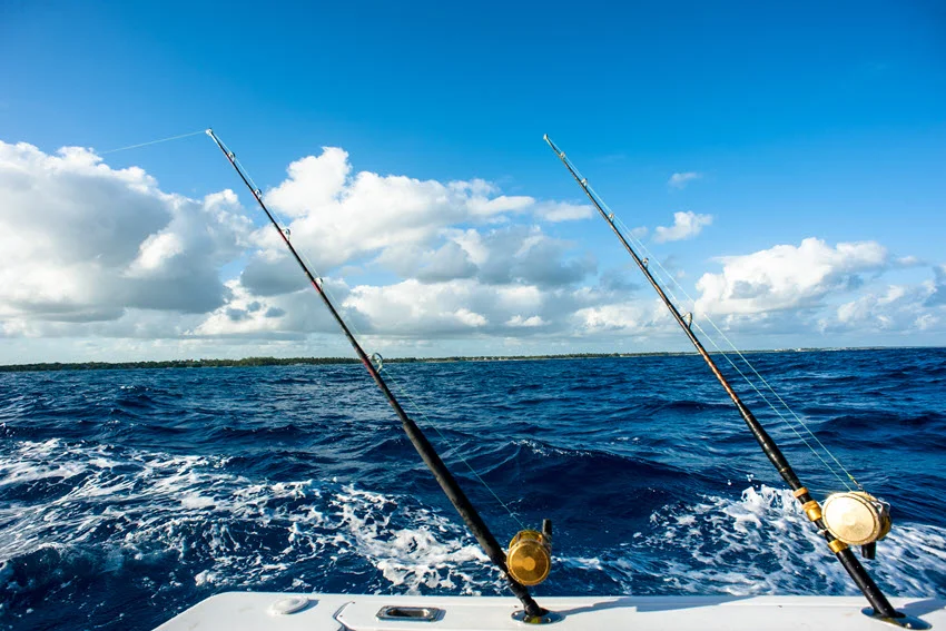 Two huge trolling rods attached to a moving boat, with the blue, open ocean and white-cloudy sky in the background.